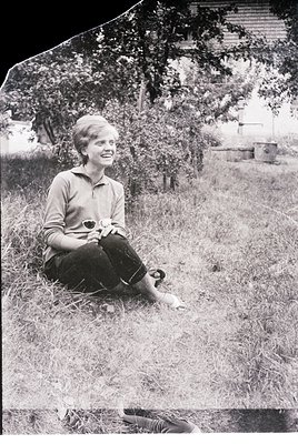 Vintage black-and-white portrait of a young woman seated in overgrown grass, holding a small object (likely a camera or lens)...