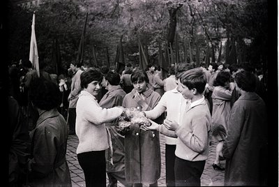 Black-and-white photo captures a public gathering in a wooded area, likely a 1960s–70s Soviet bloc country. A group exchanges...