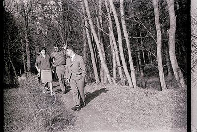 Three men in mid-20th-century attire—one in a suit, another in a sweater, and a third in casual pants—walk a narrow forest pa...