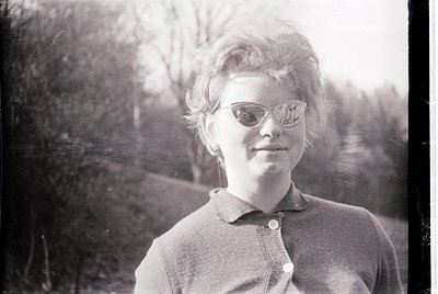 Vintage black-and-white portrait of a young woman in a short-sleeved button-up shirt, standing outdoors with blurred greenery...