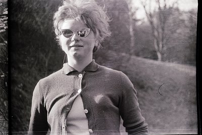 Black-and-white portrait of a person with voluminous, teased hair and round glasses, wearing a buttoned sweater over a collar...