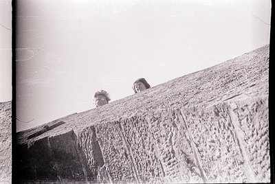 Vintage black-and-white photo of two individuals peering over a rough-textured stone wall, likely part of a historic or indus...