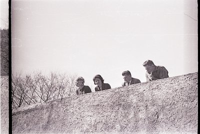 Four individuals peering over a rocky cliffside, mid-20th century black-and-white photography. Formal attire suggests a schoo...