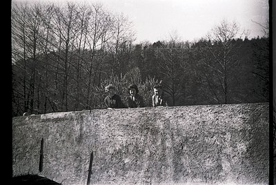 Three individuals pose on a concrete ledge, leaning against a rugged stone wall in a forested area. Mid-20th century attire s...