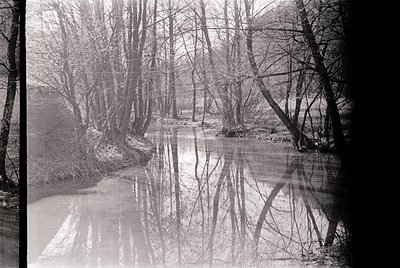 Vintage black-and-white photograph of a serene, narrow river flanked by leafless trees, creating mirrored reflections. Overca...