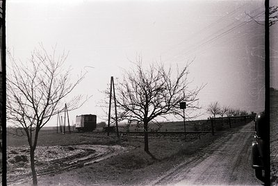 Black-and-white rural scene featuring a lone car parked on a dirt road beside a railway track. Bare winter trees line both si...