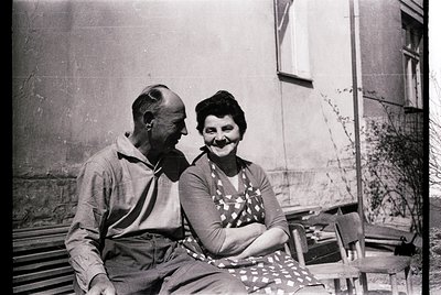 Mid-20th century black-and-white portrait of an elderly couple sharing a smile outdoors. Man wears a striped polo shirt; woma...