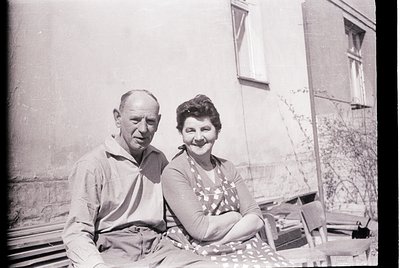 Mid-20th century black-and-white portrait of a man and woman seated on a wooden bench outside. The man wears a short-sleeve b...