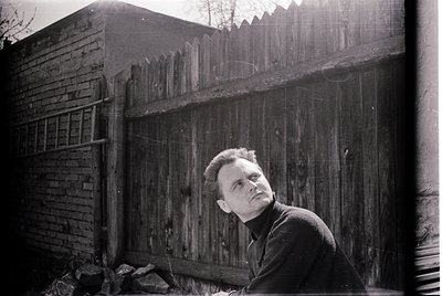Mid-century man leans against weathered brick wall behind rustic wooden fence. Classic 1950s–1960s street portrait style with...