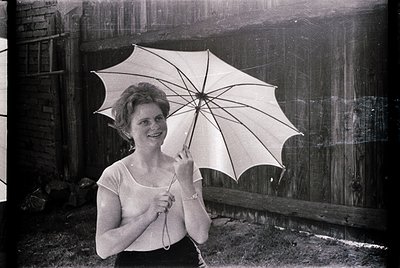 Vintage black-and-white portrait of a woman holding a large, open parasol in an outdoor setting. She wears a sleeveless dress...