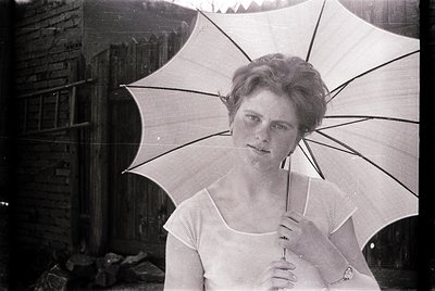 Vintage black-and-white portrait of a woman holding a large, open parasol. She wears a high-necked, sleeveless dress with a s...