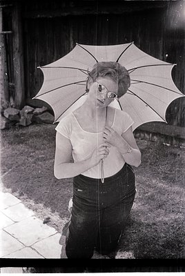 Mid-century woman in a sleek, rounded-neck blouse and high-waisted jeans holds a vintage parasol. Rustic wooden fence and cob...