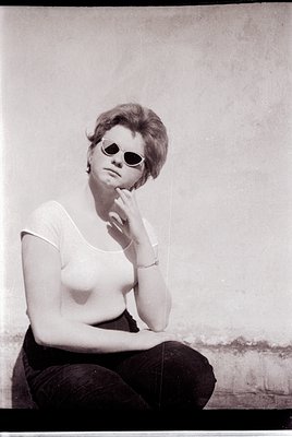 Portrait of a woman in mid-20th century fashion—sitting against a plain wall, wearing a sleeveless top, dark trousers, and ro...