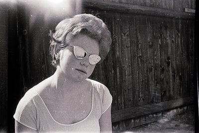 Black-and-white portrait of a woman in mid-20th century fashion—rounded sunglasses, short bob haircut, and striped blouse—sta...