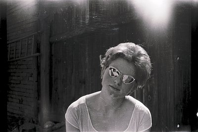 Portrait of a woman in 1960s-style round sunglasses, standing indoors near a wooden door with light streaming through. She we...