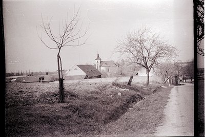 Vintage black-and-white rural scene featuring a paved road bordered by leafless trees and a low stone embankment. Distant far...