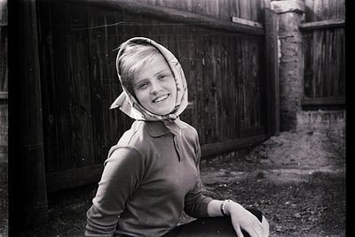 Mid-20th century black-and-white portrait of a young woman in a headscarf and long-sleeve sweater, leaning on a rustic wooden...
