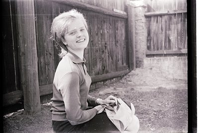 Young girl in a vintage sweater leans against a weathered wooden fence, holding a small stuffed animal. Mid-20th century urba...