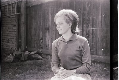 Mid-century black-and-white portrait of a young woman seated outdoors, 1950s–1960s. She wears a collared sweater and sits cro...