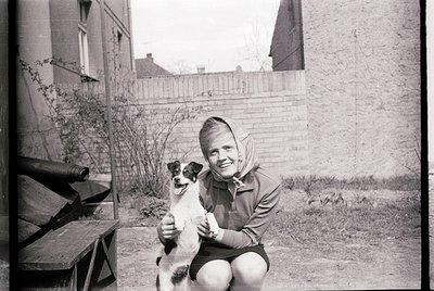 Black-and-white candid of a woman kneeling beside a small terrier-type dog in an urban backyard, mid-20th century. She wears ...