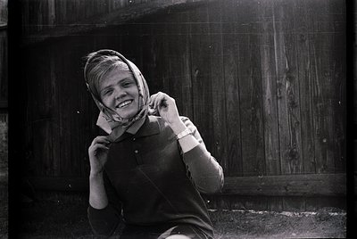 Black-and-white portrait of a young woman in a sweater and headscarf, adjusting a choker necklace. Wooden door and rustic wal...