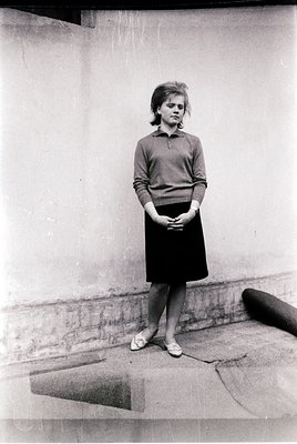 Young woman in mid-20th-century streetwear—brown sweater, knee-length skirt, and flat shoes—poses against a plain concrete wa...