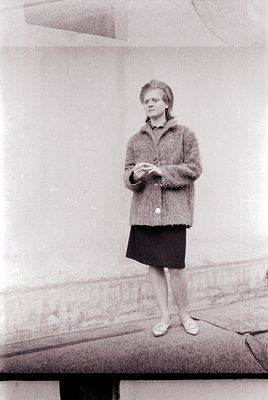 Portrait of a woman in mid-20th-century fashion, standing on a ledge against a plain wall. She wears a textured cardigan, kne...