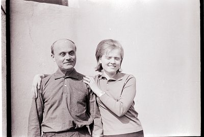 Black-and-white portrait of a man and woman posing closely against a plain wall. The man wears a striped button-down shirt wi...