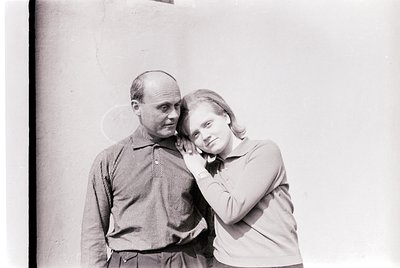 Black-and-white portrait of a man and woman in close embrace, leaning against a plain wall. The man wears a striped button-up...