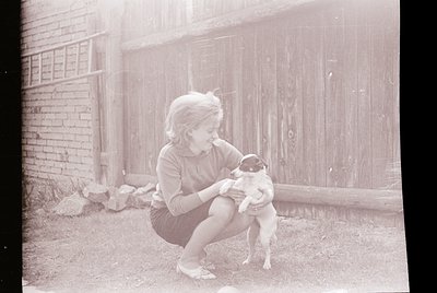 Vintage black-and-white photo of a young girl squatting outdoors, holding a small dog. Brick wall and wooden fence in backgro...