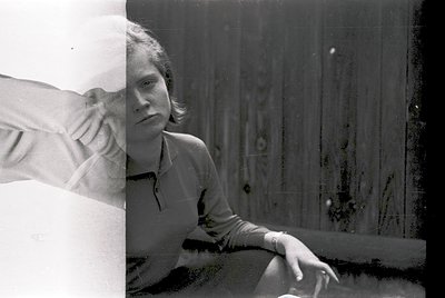 Vintage black-and-white portrait of a person seated indoors, leaning on a table with a blurred arm resting on it. The individ...