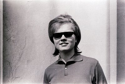 Portrait of a young woman in 1960s-style sunglasses and short, layered hair, wearing a polo shirt. Plain concrete wall backgr...