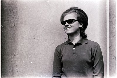 Black-and-white portrait of a person in 1960s-style sunglasses and a polo shirt, leaning against a rough-textured wall. The h...