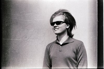Black-and-white portrait of a person with voluminous 1960s-style hair and sunglasses, wearing a collared polo shirt. The cand...