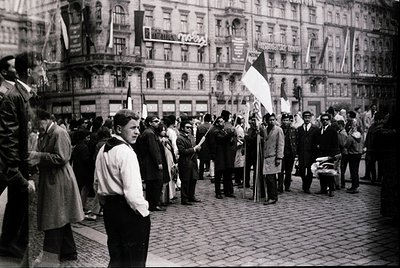 Crowd of men in 1950s-60s street protest, holding flags with red/yellow/white stripes. Architectural backdrop of ornate Europ...