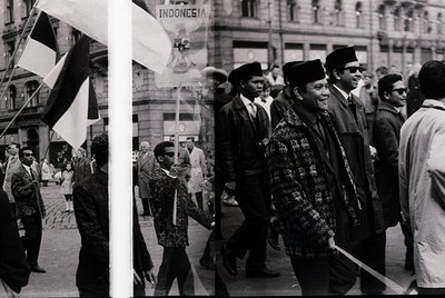 Black-and-white street scene featuring Indonesian national flags and crowds in 1960s Jakarta. Central figures wear traditiona...