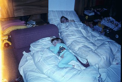 Two individuals sleep on a disheveled bed in a dimly lit room, likely a 1970s-80s domestic interior. The person in the foregr...