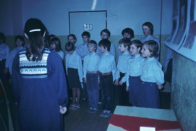 A group of children in coordinated 1970s-style school uniforms (light blue shirts, dark skirts/pants) stand in a semicircle, ...