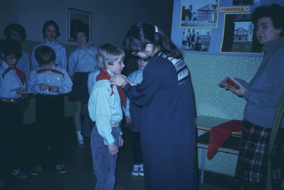 Scout ceremony featuring young boy receiving a neckerchief from adult leader in 1970s indoor setting. Group of children in ma...