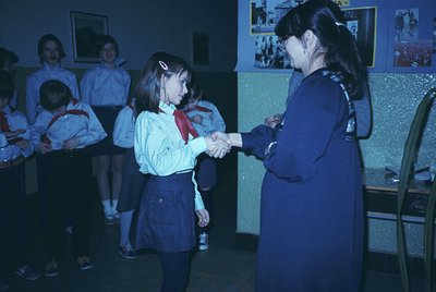 1970s-era school ceremony: young girl in red-and-white scarf uniform shakes hands with adult in blue uniform. Indoor setting ...