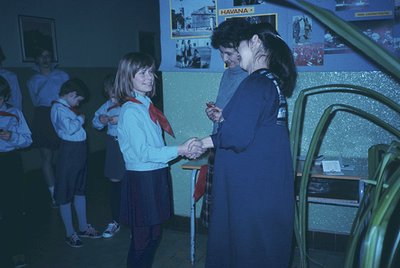 A 1970s indoor school scene: two girls in matching uniforms (light blue blouses, red scarves, dark skirts) shake hands with a...