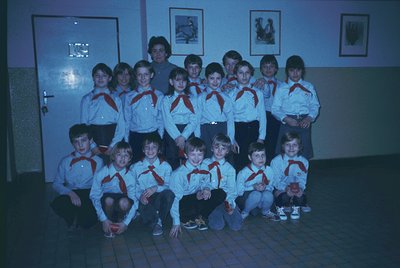 Group photo of youth in scout uniforms (red scarves, white shirts) posed indoors, likely 1970s–1980s. Indoor setting with fra...