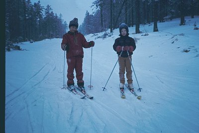Two children pose on groomed ski slopes in vintage ski gear, surrounded by dense evergreen forest. Likely mid-20th century al...