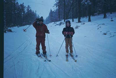 Two children pose on a groomed alpine slope, wearing vintage ski gear—wide skis, bright yellow bindings, and striped ski pole...