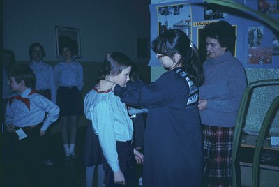 Scout uniform ceremony in a dimly lit hall, featuring a leader (right) pinning a badge on a girl’s uniform. Group of children...