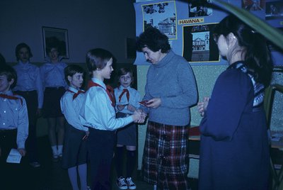 Scout leader in plaid skirt and sweater hands a certificate to a boy in scout uniform (1970s style) during an indoor ceremony...