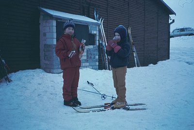 Two children pose with vintage skis in snowy setting, likely mid-20th century. Boy on left wears maroon ski gear with helmet;...