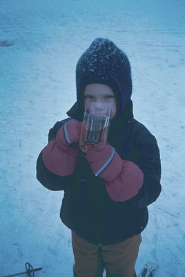 Young child in winter attire—knit beanie, padded mittens, and brown pants—holds a clear glass filled with liquid, likely hot ...