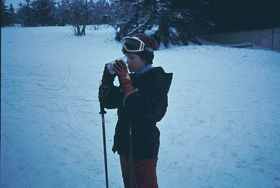 Winter skier pauses mid-descent, holding a camera to capture snowy alpine scenery. Mid-20th century gear: goggles, scarf, and...
