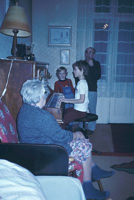 Vintage indoor scene featuring a seated elderly woman playing a piano with three children and an adult male observing. Mid-ce...